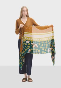Mønstret flerfarvet tørklæde med blomsterdesign, grønne og sennepsfarvede sektioner, glat tekstur og frynsekanter, holdt af en model.