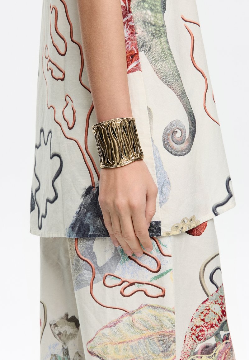 Amplia pulsera de oro con un diseño texturizado y ondulado, llevada en una mano. El fondo presenta una tela clara con un patrón suelto.