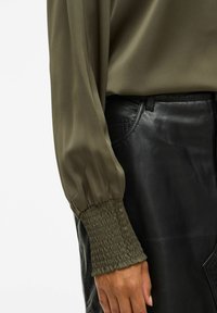 Getailleerde olijfgroene blouse met manchet en elastische pols, gecombineerd met hoog gewaarmerkte zwarte leren broek.