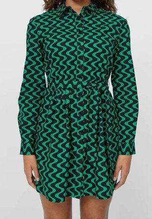 Woman wearing a long-sleeve, button-up dress with a green and black wavy pattern and a cinched waist.