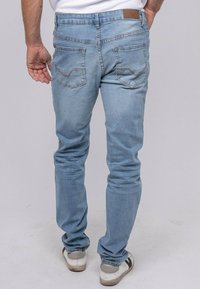 Un homme portant un jean bleu clair et des baskets blanches se tient de dos, visible contre un fond uni.