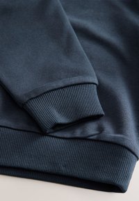 Tissu de sweat-shirt bleu foncé avec poignets côtelés. Texture lisse avec une coupe ajustée. Détail de couture visible le long des coutures.