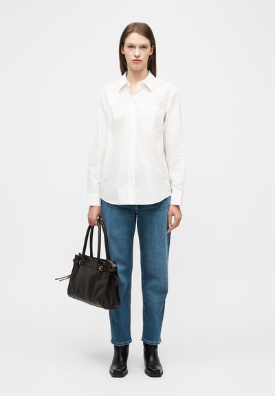 Femme vêtue d'une chemise blanche à boutons, d'un jean bleu et de bottes noires, tenant un sac à main noir sur un fond blanc uni.