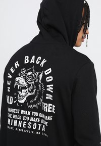 Black hoodie with a graphic print featuring a wolf and text: "NEVER BACK DOWN," "WILD & FREE," and "MINNESOTA." Text in white on back.