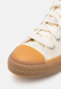Canvas sneaker with creamy upper, orange rubber toe cap, white laces, and grey metal eyelets; features textured gum rubber sole.