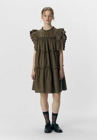 Braun gestuftes Kleid mit Rüschensleeves, rundem Ausschnitt und weichem Stoff. Tragen mit schwarzen Socken und Schuhen, vor einem schlichten Hintergrund.