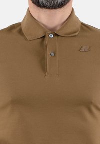 Polo marrone realizzato in tessuto testurizzato, con colletto e pattina a tre bottoni. Include un piccolo logo sulla parte del petto.