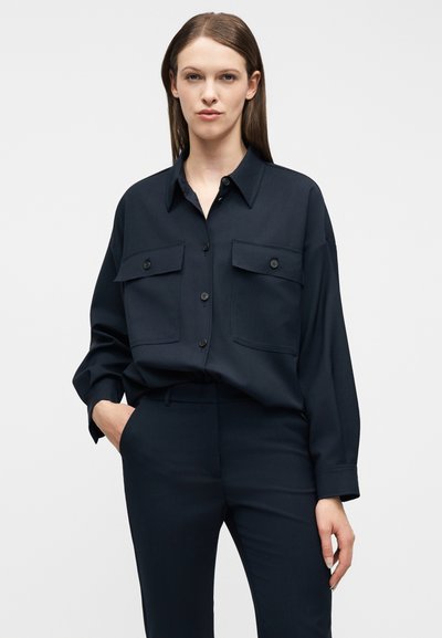 Γυναίκα που φοράει ένα σκούρο μπλε πουκάμισο με κουμπιά και δύο τσέπες στο στήθος, καθώς και παντελόνι με ψηλή μέση που ταιριάζει, στέκεται με το ένα χέρι στην τσέπη.