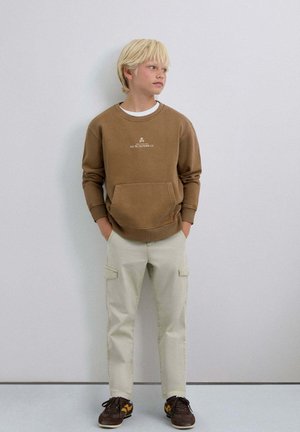 Sudadera marrón con bolsillo delantero y logo blanco, combinada con pantalones cargo claros y zapatos marrones con detalles amarillos.