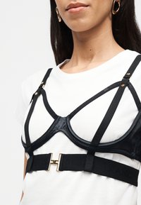 Γυναίκα που φοράει μαύρο σουτιέν-ιμάντα πάνω από ένα απλό λευκό μπλουζάκι, με χρυσές μεταλλικές λεπτομέρειες στους ιμάντες και  τη ζώνη.