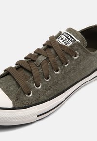Baskets Converse basses vert olive avec lacets marron, œillets en métal, bout en caoutchouc blanc et bande noire sur une semelle blanche.