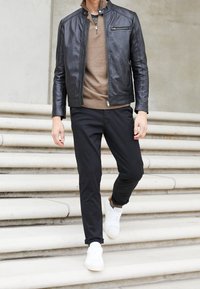 Black leather jacket, brown sweater, and black pants, paired with white sneakers; model walking down white stone steps.