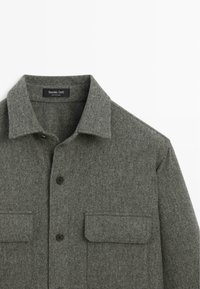 Grå uldskjortejakke med en spredt krave, sort knaplukning og to forlommer med lapper. Blødt, tekstureret stof med et struktureret design.