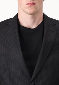 Blazer nero a righe con revers a punta, chiusura a un bottone e taschino sul petto, indossato sopra una t-shirt nera semplice.