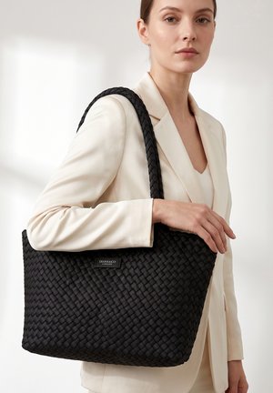 Donna con blazer color crema che tiene una grande borsa tote nera intrecciata con spalline e etichetta del marchio visibile.