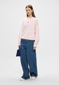 Różowy sweter z opadającymi ramionami, zestawiony z oversize'owymi niebieskimi dżinsami w wzory. Dodatki obejmują małą niebieską torebkę.