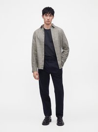 Jeune homme debout, portant des chaussures noires, pantalon foncé, chemise noire et veste beige à carreaux ouverte, main gauche dans la poche sur fond blanc.