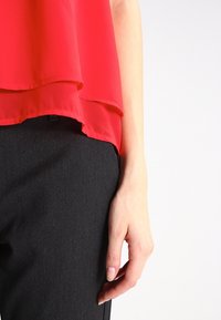Blusa rossa a strati sopra pantaloni grigi scuri con una texture di stoffa liscia. La blusa presenta un design fluido e una vestibilità morbida.
