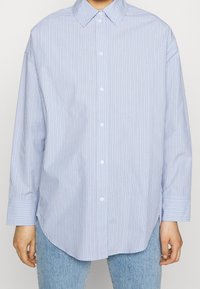 Light blue long-sleeve button-up shirt with vertical white and dark blue stripes, worn untucked over light blue jeans.