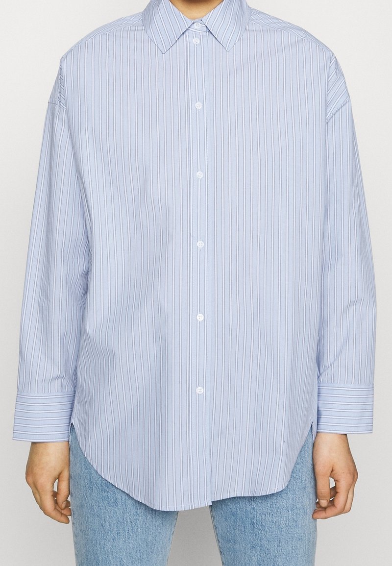 Light blue long-sleeve button-up shirt with vertical white and dark blue stripes, worn untucked over light blue jeans.