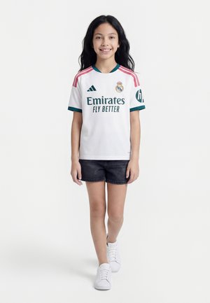 Fille aux longs cheveux portant un maillot de football blanc du Real Madrid, un short noir et des baskets blanches, marchant vers la caméra avec un sourire.