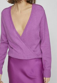 Růžový pletený svetr s výstřihem do V a dlouhými rukávy, s designem zavinování. Nošený s odpovídající fialovou saténovou sukní, která má hladkou texturu.