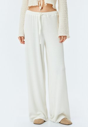 Pantalones blancos de pierna ancha, hechos de un tejido texturizado, con una cinturilla ajustable mediante cordón, llevados con unos zapatos beiges tipo slip-on.