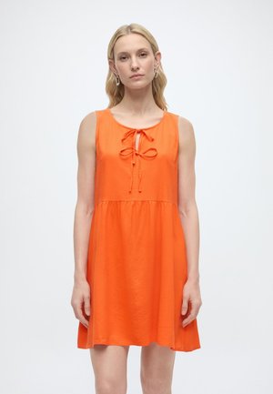 Kvinde med lyst hår iført ærmeløs, knaldrød kjole med bindebånd foran, stående mod en ensfarvet lys baggrund.