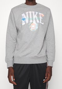 Person som bär grå Nike-tröja med färgstarkt logo och text, kombinerad med svarta Nike-shorts med vit swoosh och sidostripes.