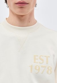 Νεαρό άτομο που φοράει φούτερ σε κρεμ απόχρωση με ριμπ κολάρο και την επιγραφή "EST 1978" σε ανοιχτό μπεζ στο στήθος.