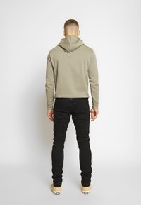 Olivově zelená mikina s kapucí na stažení, černé slim fit kalhoty a béžové boty. Hladká textura, minimální detaily.