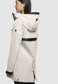 Veste imperméable blanche avec capuche, accents noirs et bandeau logo au dos. Elle présente des fermetures à pression et des poignets texturés.