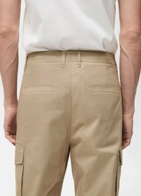 Beige Cargo-Hosen mit glatter Textur, ausgestattet mit zwei Gesäßtaschen und einer Seitentasche, kombiniert mit einem eng anliegenden weißen Hemd.