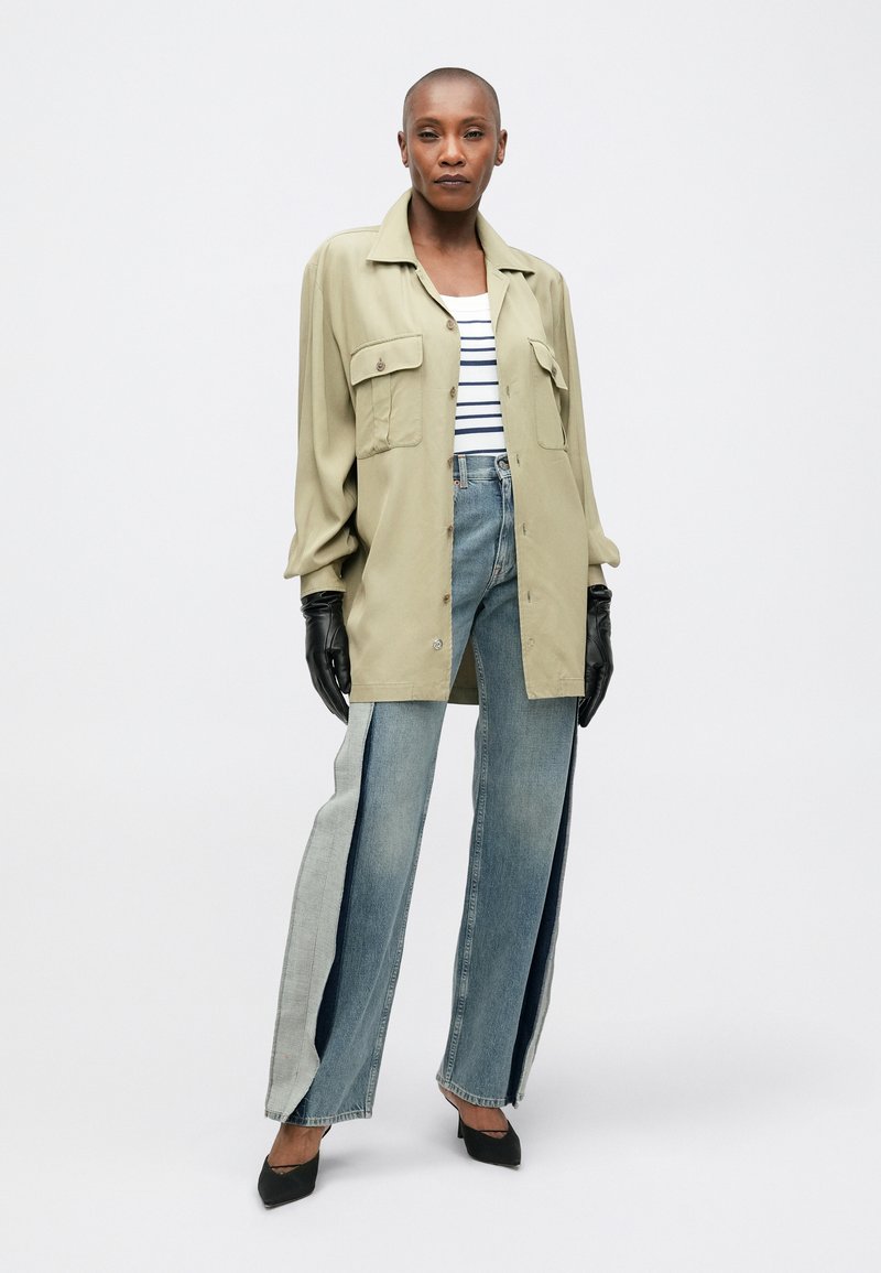 Ljusgrön överdimensionerad skjortjacka med två bröstfickor, parat med denimjeans med en delad design i olika nyanser av blått. Svarta handskar.