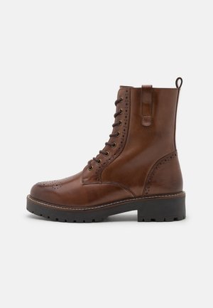 Brązowy skórzany botek za kostkę z wiązaniem na sznurówki, zdobieniami typu brogue, grubą czarną gumową podeszwą i pętelką do ściągania na białym tle.