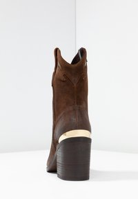 Hnědé semišové kotníkové boty s nízkým dřevěným podpatkem, které mají zlatý metalický akcent na podpatku a dekorativní výřezy po stranách.