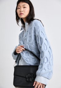 Gaiši zils adīts džemperis ar cilpu rakstu, uzvilkts virs gaiši zilas krekla. Melna ādas rokassoma ar izspiestu logo un siksnu.