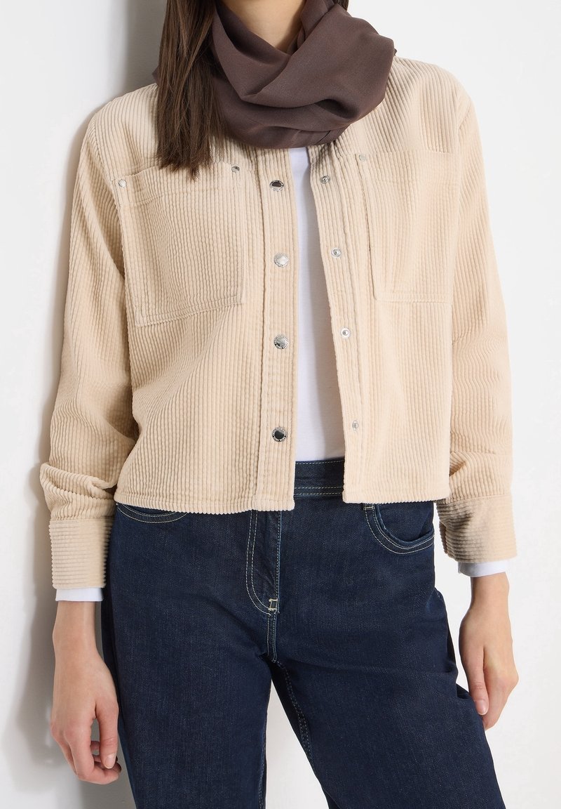 Veste-chemise en velours côtelé beige boutonnée portée sur un top blanc, associée à un jean bleu foncé et une écharpe marron autour du cou.