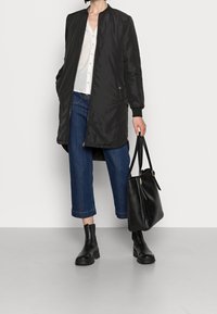 Personne portant une longue veste noire, une chemise blanche boutonnée, un jean bleu court, des bottes noires, tenant un sac fourre-tout en cuir noir.