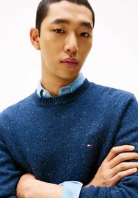 Blauer Strickpullover mit rundem Ausschnitt und gesprenkelter Textur. Getragen über einem hellblauen Hemd mit Kragen. Kleines Logo auf der Brust.