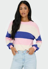 Femme aux longs cheveux bruns portant un pull rayé rose, crème et bleu et un jean bleu clair, les mains dans les poches.