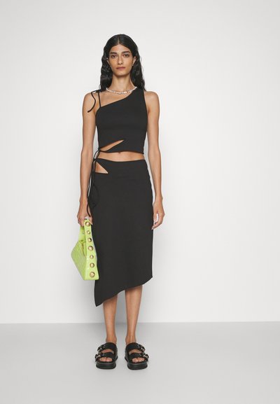 Top court noir avec un design asymétrique et des découpes, associé à une jupe noire taille haute avec un ourlet asymétrique. Le modèle tient un sac vert vif.