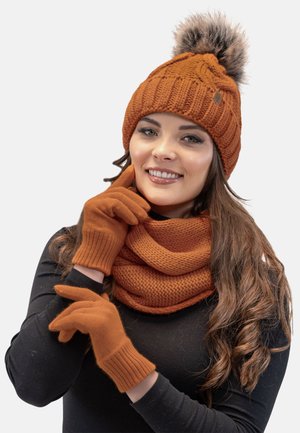 Un bonnet orange en tricot avec un pompon en fausse fourrure, des gants assortis et une écharpe infinity texturée présentent un design cohérent et une palette de couleurs chaudes.