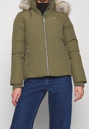 Person wearing an olive green puffer jacket with fur-lined hood and blue jeans, standing against a plain background.