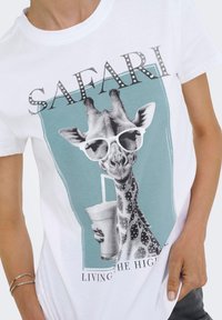 Camiseta blanca de algodón con un gráfico de una jirafa usando gafas de sol, sosteniendo una bebida, con el texto “SAFARI” arriba y “VIVIENDO LA VIDA ALTA” abajo.