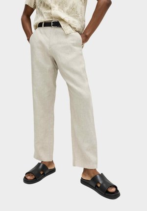 Man draagt beige linnen broek, lichtbruine blouse met patroon, zwarte riem en zwarte open teen slippers, waarbij zijn voeten gedeeltelijk zichtbaar zijn.