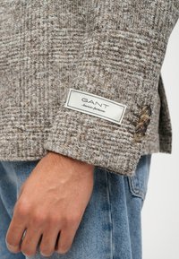 Brown tweed blazer with a textured pattern, featuring a GANT label on the sleeve and four button accents on the cuff. Denim visible underneath.