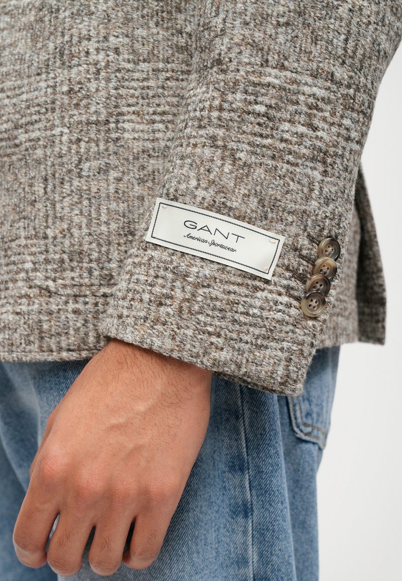 Bruine tweed blazer met een gestructureerd patroon, voorzien van een GANT-label op de mouw en vier knoopaccenten op de manchetten. Spijkerbroek zichtbaar eronder.