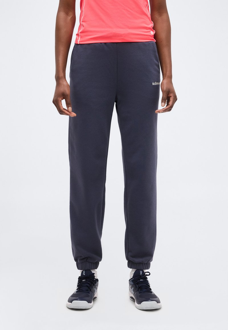 Persoană purtând pantaloni de trening bleumarin cu manșete elastice, adidași negri și albi, și un top roșu fără mâneci pe un fundal simplu.