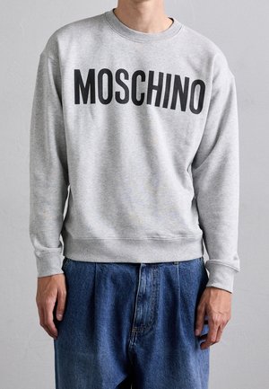 Person iført en lysegrå sweatshirt med "MOSCHINO" i store sorte bogstaver over brystet og løse blå jeans.